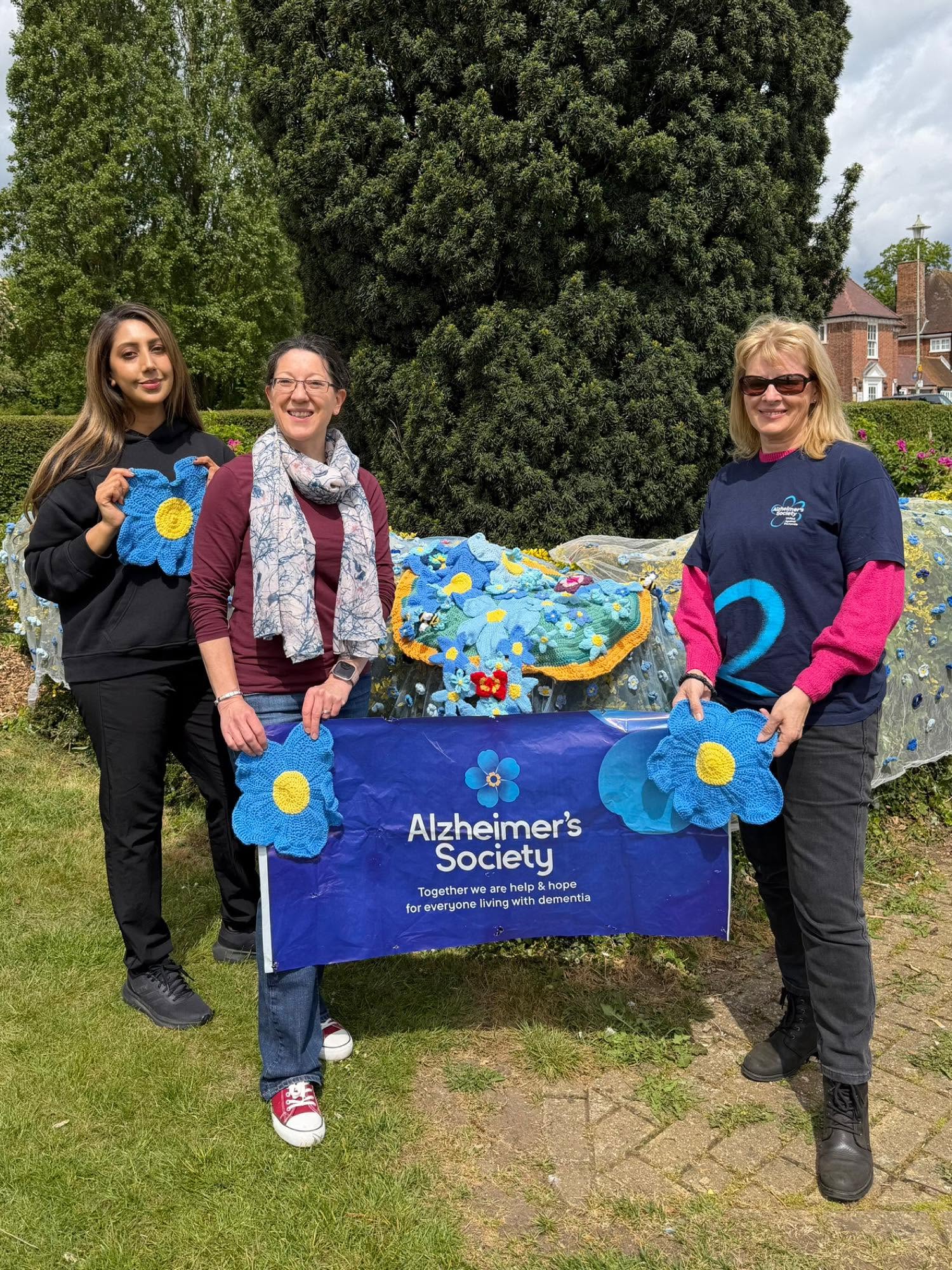 Three people holding blue knitted flowers and an Alzheimer's Society banner outdoors